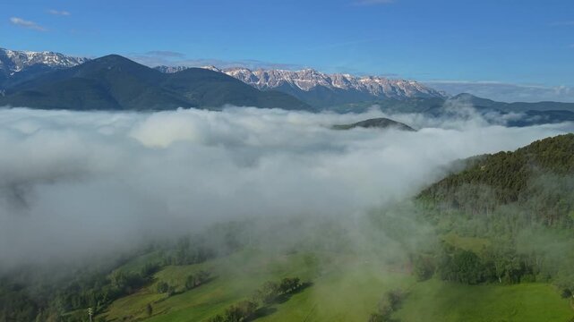 Mountain peaks rising above sea of clouds at sunrise in Catalonia, Spain, aerial drone view of mist covered valley, green meadows and forested hills 