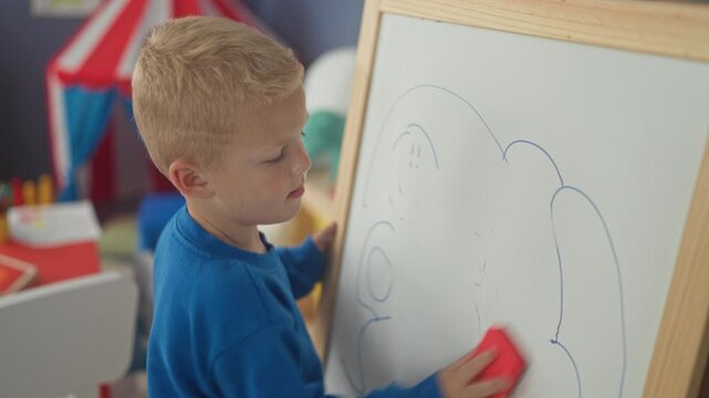 Boy wiping whiteboard with red eraser, hand rubbing drawn cloud on board in building; concentration learning.