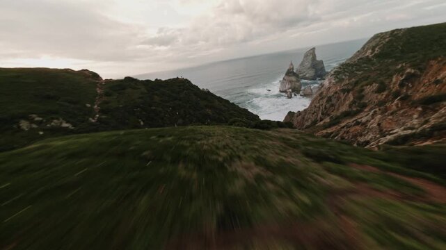 Fast FPV dive from rocky green cliff toward Ursa Beach coastline during sunset