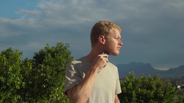 Blonde man using an electric trimmer on his jawline and neck with eyes closed. Green trees and mountains under a blue sky with clouds in the background. Static medium shot.