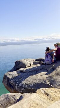 Vertical. Mom and kids sit on boulder on sea cliff overlooking ocean at Robberg