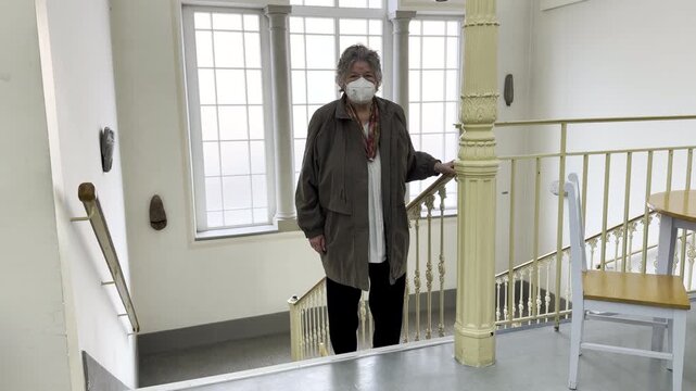 An elderly woman is walking up the stairs in an office building; she is wearing a mask, static shot