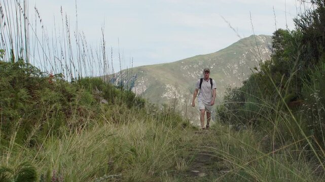 Solo male hiker wearing a cap and backpack treks forward along a narrow path through tall grass, with rolling green mountains visible in the background. Static eye level wide view.
