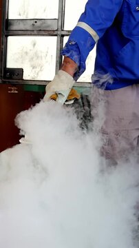 A worker wearing protective gloves and blue uniform handles a container emitting thick white nitrogen vapor.