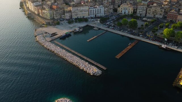 Slow panoramic drone reveal of Corfu Old Town at sunrise. Promenade visible along golden waterfront. Clear sky. Greece aerial footage