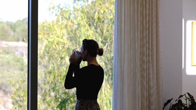 Woman sips coffee while looking out of a large window in her house