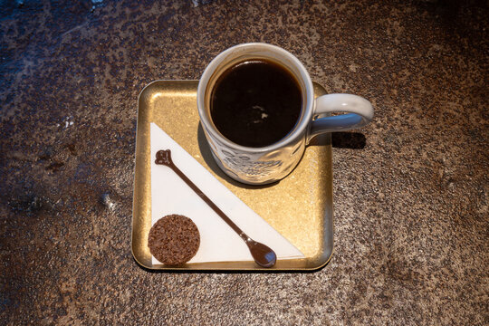 Top-down view of a steaming cup of black coffee in a patterned ceramic mug on a gold tray with a chocolate cookie and decorative spoon, suggesting a cozy morning ritual and simple comfort.