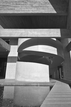 Dramatic black and white image of a modern concrete spiral staircase framed by geometric minimal architecture, strong light and shadow, and reflective water creating a sculptural, contemplative