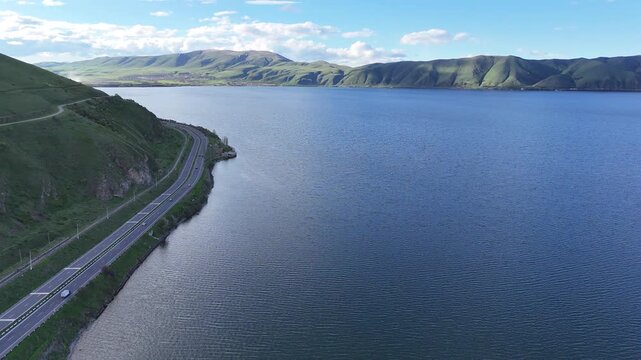 4K Drone Aerial View of Scenic Mountain Road Along Large Blue Lake With Green Hills and Natural Landscape.