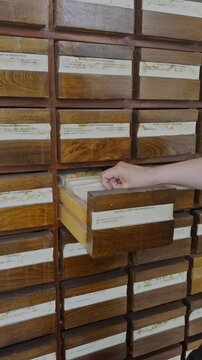 Woman's hand searching through index cards in a wooden library drawer. Vintage archives, research, rustic textures, and close-up detail.
