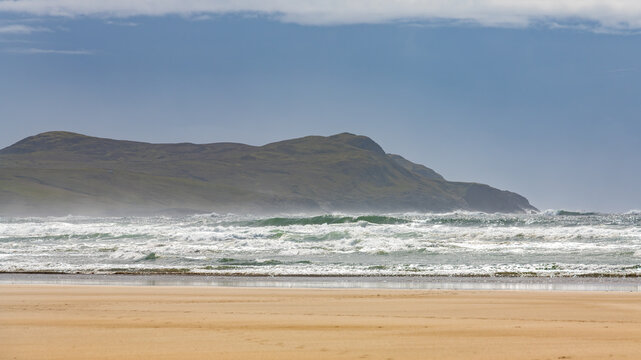 Scenic view of Machir Bay Beach on the Isle of Islay in Scotland, featuring powerful ocean waves rolling onto the sandy shore with rugged cliffs in the background. 