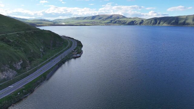 4K Drone Aerial View of Scenic Mountain Road Along Large Blue Lake With Green Hills and Natural Landscape.