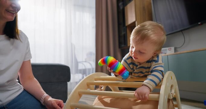 A young toddler engaging in sensory play with a colorful stretchy toy while sitting on a wooden climbing structure in a cozy living room environment during daytime