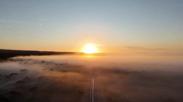 Aerial drone ascent pulling back above morning fog toward the red sunrise over a straight road in Australia  