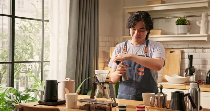 Young Asian home barista in apron using manual hand grinder to grind coffee beans. Smiling gen-z male preparing fresh coffee in kitchen. Slow life, specialty coffee hobby, and morning routine concept.