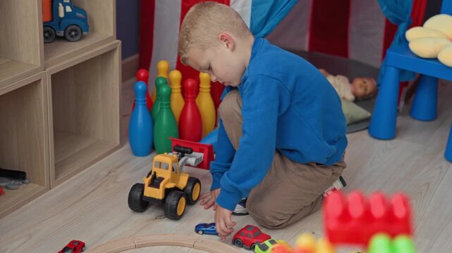 Boy kneeling and reaching for toy cars and a yellow loader near wooden cubed shelving and colorful bowling pins inside a playroom building; curiosity imagination.