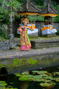 Young Balinese legong dancer in gold headdress and green batik costume standing on mossy stone steps at temple pond