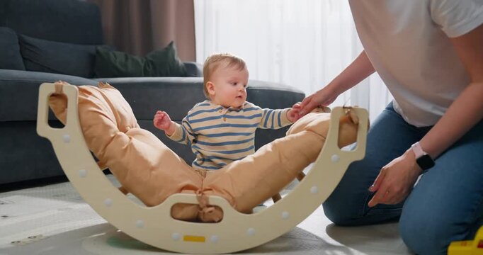 A young baby explores a cushioned wooden rocker toy with the assistance of an adult, engaging in early play and motor skill development in a cozy living room environment.