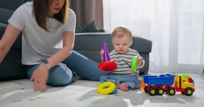 Young mother playing and interacting with her toddler son on the floor using colorful stacking rings and toy truck in a bright, cozy living room environment