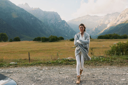 A young woman walks along a gravel road in a mountain valley, wrapped in a light cardigan, conveying a calm outdoor lifestyle and solo exploration under open skies.