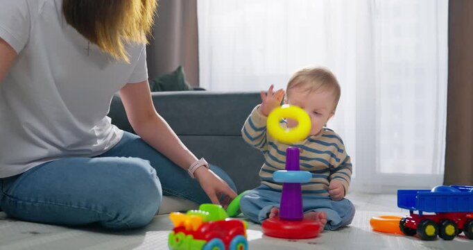 A toddler in a striped outfit plays with colorful plastic stacking rings while sitting on the floor near a woman engaging in interactive play with various vibrant toys in a cozy living room setting