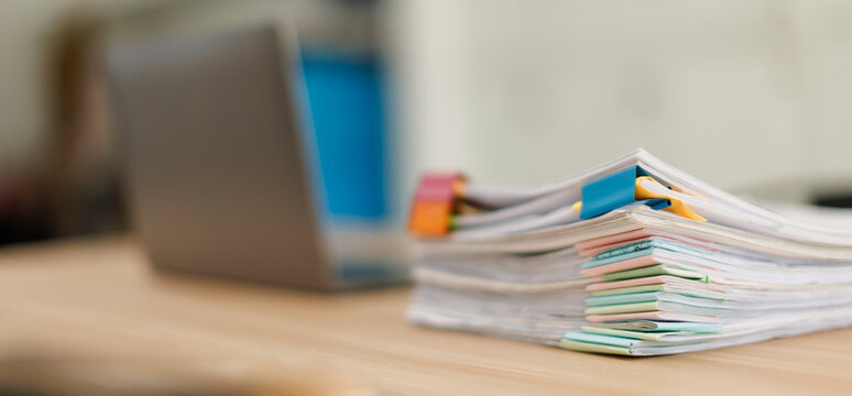 Stack of document files, paperwork, and reports organized with binder clips on a desk next to a blurred laptop, representing business management and data organization