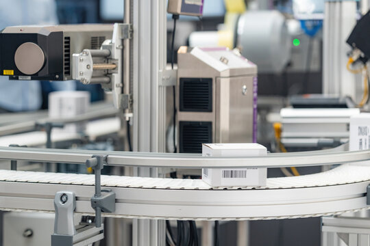 Automated industrial packaging and labeling system processes small white boxes on a curved conveyor belt, captured during a live demonstration at an indoor manufacturing technology exhibition.