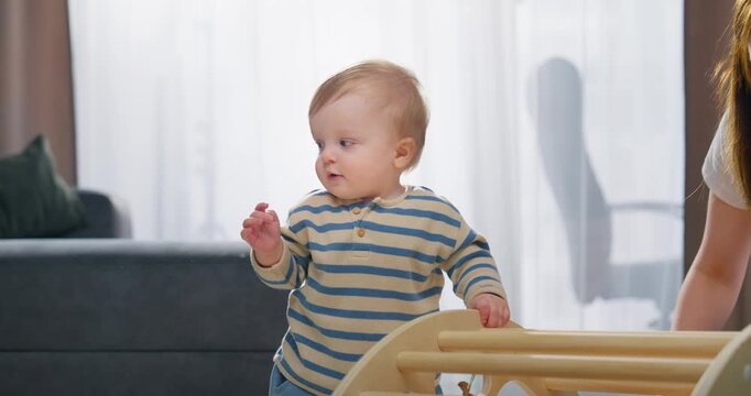 A young toddler learning to stand and walk with the supportive guidance of an adult in a cozy indoor setting featuring wooden climbing equipment and soft natural lighting