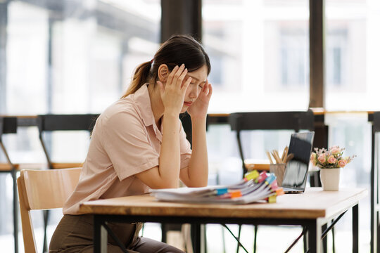 Stressed young asian business woman suffering from a severe headache or migraine while working at her office desk. Overwhelmed by work pressure. Fatigue. And burnout