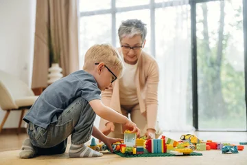 Fotobehang Lego Young boy with glasses playing with colorful building blocks on the floor while grandmother watches and assists in a bright living room with large windows  © standret