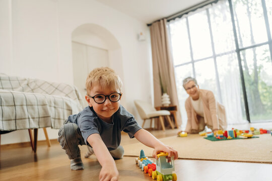 Young boy with glasses playing with toy train on wooden floor in bright living room, grandmother sitting on sofa in the background, large windows letting in natural light