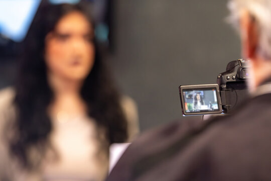 Live media interview with a young woman recorded at an indoor trade fair, with professional video camera capturing the moment from behind the cameraman during an active press session.