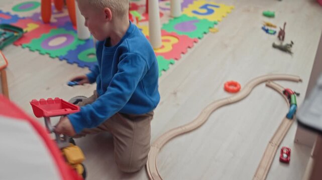Boy kneeling plays with toy dump truck and red shovel on foam number mat in building; playful curiosity.