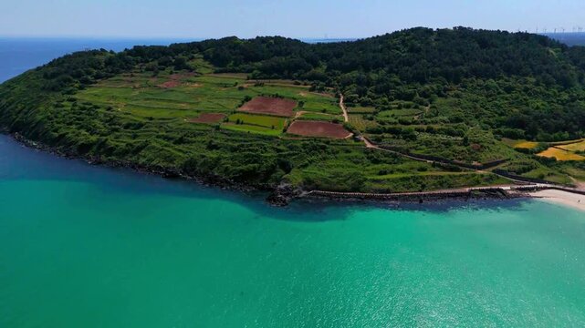 Aerial vista of a green volcanic cape covered with patchwork farm fields surrounded by deep blue and emerald sea, Hamdeok, Jeju Island, South Korea