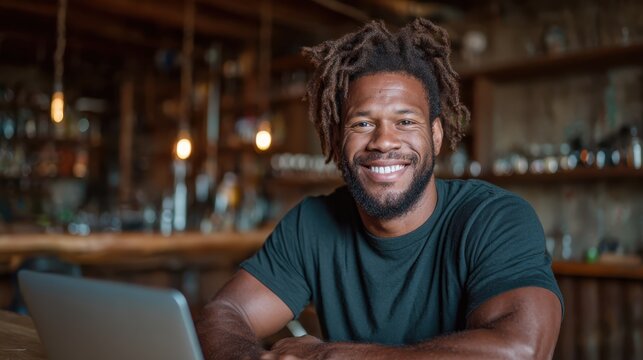 A smiling man with dreadlocks, sitting at a cafe with a laptop and warm ambiance, embodying a relaxed and personal atmosphere while striving for creativity and productivity.