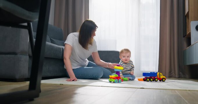 Mother interacting with her baby boy playing with colorful toy vehicles and stacking blocks on the floor of a bright living room during daytime, fostering early childhood development and bonding.