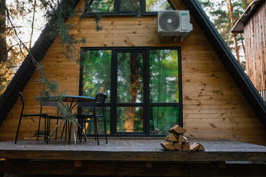 A-frame log chalet house in the forest with a natural backdrop. Summer getaway in a wooden cottage. Outdoor terrace, seating area, firewood