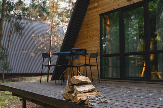 A-frame log chalet house in the forest with a natural backdrop. Summer getaway in a wooden cottage. Outdoor terrace, seating area, firewood