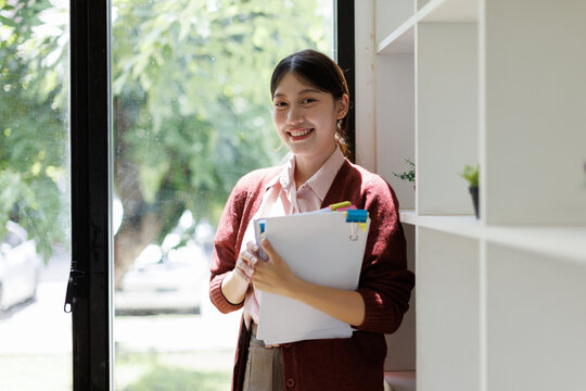 Young asian woman smiling confidently in a bright office by a window, holding documents and paper clips&mdash;symbol of student ambition, professional growth and organized career focus