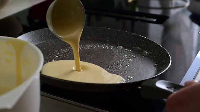 Slow motion close up of homemade crepe batter being poured into a hot frying pan in a private kitchen. Traditional cooking process, breakfast preparation and home baking.