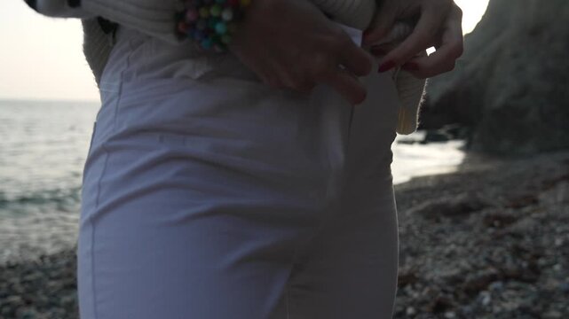 Woman zipping up white pants, fastening the zipper and button while standing on a rocky beach during sunset.