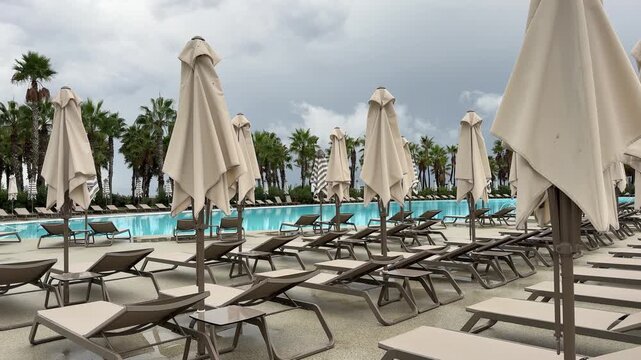 Sun loungers and closed umbrellas by resort pool on cloudy day