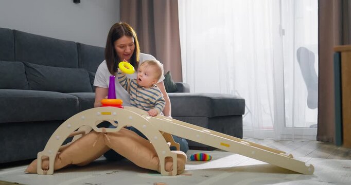 Mother and toddler playing together with a wooden toy ramp and ring stacking set in a bright, cozy living room featuring soft carpet and natural daylight from large windows