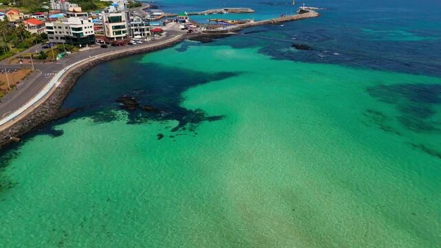Aerial view of crystal-clear gradient turquoise water over a rocky reef beside a coastal road and a small fishing harbor, Hamdeok, Jeju Island, South Korea