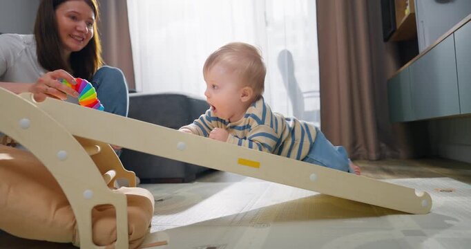 Mother engaging with her baby playing on a wooden climbing ramp in a cozy living room setting, promoting early childhood development through interactive play and bonding time
