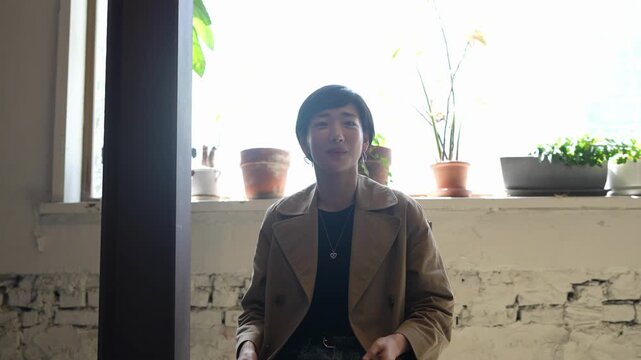 Soft light fills a cozy room with plants. Woman smiles, gesturing with open hands. Her coat and calm expression invite connection. Ideal for lifestyle, wellness, or creative content