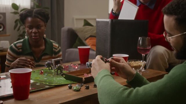 Tilt down shot of group of friends listening to game master behind screen and eating snacks while playing tabletop RPG game at home in cozy living room