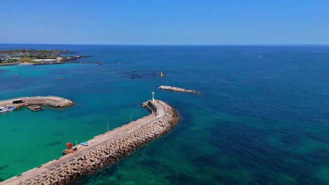 Aerial view of a breakwater with a lighthouse and a small fishing harbor between turquoise and deep blue sea, Hamdeok, Jeju Island, South Korea