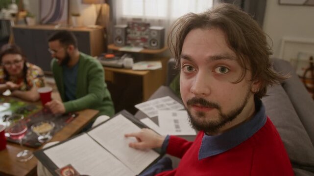 High angle close up portrait shot of young bearded game master looking up at camera while reading script for RPG tabletop game with friends in cozy living room