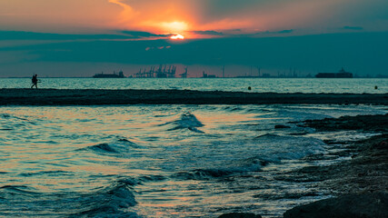 Obraz premium Man walking along beach during industrial port sunset